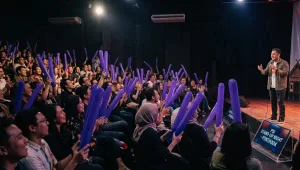 Penonton mengangkat balon tepuk berwarna ungu saat menyaksikan acara komedi di dalam ruangan.